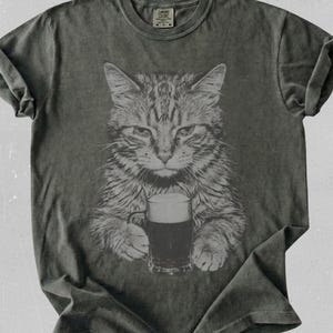 Puede incluir: Una camiseta de manga corta en color gris carbón con un estampado gráfico de un gato atigrado sosteniendo una jarra de cerveza oscura. La prenda tiene cuello redondo y un acabado lavado. La etiqueta interior muestra el texto Comfort Colors.