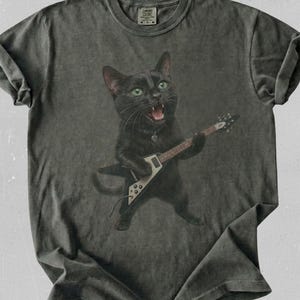 Peut inclure: Un t-shirt gris anthracite &agrave; manches courtes orn&eacute; d'un motif repr&eacute;sentant un chat noir aux yeux verts jouant d'une guitare &eacute;lectrique blanche et noire. Le chat a la gueule ouverte comme s'il chantait. Le t-shirt a un col rond et un aspect d&eacute;lav&eacute;.