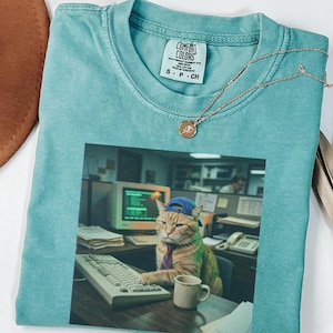 Può includere: Una t-shirt verde menta gessato con una stampa grafica di un gatto soriano arancione che indossa un berretto da baseball blu e una cravatta, seduto a una scrivania con un computer, una tastiera e una tazza. La maglietta ha l'etichetta Comfort Colors.