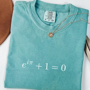 Puede incluir: Una camiseta de color verde azulado con la ecuación e^iπ + 1 = 0 impresa en blanco. Un collar dorado con un colgante circular descansa sobre la camiseta. La camiseta está doblada y tiene una etiqueta que dice "Comfort Colors".