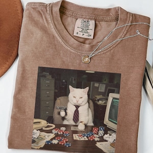 Puede incluir: Una camiseta color espresso con un estampado gráfico de un gato blanco con corbata, sentado en un escritorio con fichas de póquer, cartas y una pistola. La camiseta presenta una etiqueta de Comfort Colors en el cuello interior.