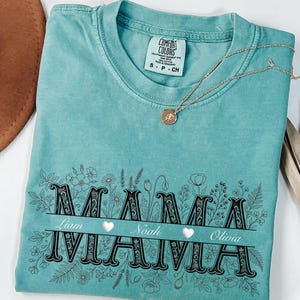 Puede incluir: Una camiseta turquesa Comfort Colors con la palabra "MAMA" en una fuente decorativa, con los nombres Liam, Noah y Olivia debajo. El diseño floral rodea el texto. Un collar dorado con un colgante descansa sobre la camisa.