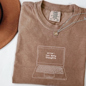 Puede incluir: Camiseta color espresso con un gráfico de portátil y el texto "error: too many thoughts". Un collar plateado con un colgante descansa sobre la camiseta. La camiseta está doblada, con un sombrero marrón en el fondo.