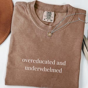 Puede incluir: Una camiseta de manga corta de color marrón con el texto "overeducated and underwhelmed" impreso en blanco en la parte delantera. La camiseta tiene cuello redondo y una etiqueta de la marca Comfort Colors en el interior. Un collar plateado descansa sobre la tela.