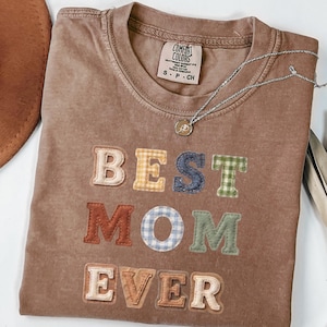 Puede incluir: Camiseta color espresso con las palabras "BEST MOM EVER" en letras coloridas estilo patchwork. Un collar plateado con un colgante descansa sobre la camisa. La camiseta tiene una etiqueta Comfort Colors.