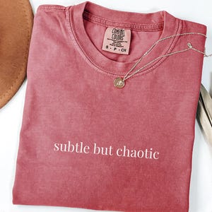 Puede incluir: Una camiseta de manga corta color carmesí con cuello redondo que presenta el texto blanco "subtle but chaotic" impreso en el pecho. La etiqueta de la marca Comfort Colors en el interior del cuello muestra la talla S P CH y 100% algodón.