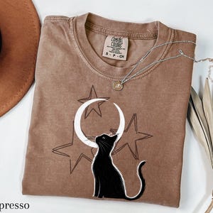 Puede incluir: Camiseta color espresso con un diseño de gato negro mirando una luna creciente y estrellas blancas. La camiseta tiene cuello redondo y una pequeña etiqueta. Un collar plateado con un colgante está drapeado sobre la camiseta.