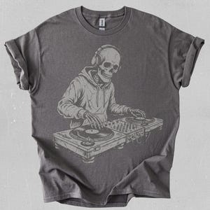 Puede incluir: Camiseta gris oscuro con una ilustración detallada de un esqueleto con auriculares y una sudadera con capucha, operando una mesa de DJ con tocadiscos. El diseño está impreso en gris claro.