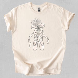 Puede incluir: Camiseta color crema con un dibujo lineal en blanco y negro de zapatillas de ballet atadas con un lazo y un ramo de flores. El diseño está centrado en la parte delantera de la camiseta, creando una estética sencilla pero elegante.