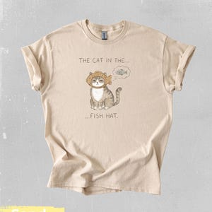 May include: Sand-colored t-shirt featuring a whimsical illustration of a cat wearing a fish hat. The text above the cat reads "THE CAT IN THE..." and below it, "...FISH HAT." The cat is gray and white, with a thought bubble showing a fish.