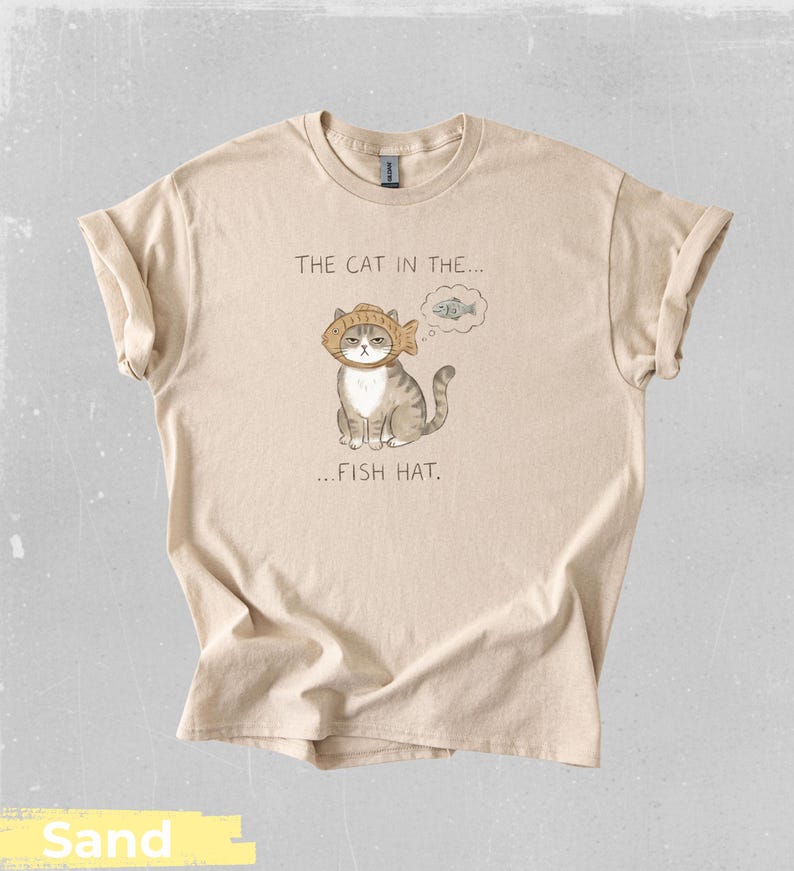 May include: Sand-colored t-shirt featuring a whimsical illustration of a cat wearing a fish hat. The text above the cat reads "THE CAT IN THE..." and below it, "...FISH HAT." The cat is gray and white, with a thought bubble showing a fish.