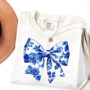 May include: Cream-colored t-shirt featuring a large blue and white floral bow design. The bow is detailed with roses and leaves. A gold necklace with a pendant and a brown hat are visible.