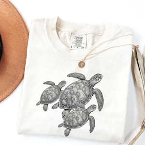 Puede incluir: Camiseta Comfort Colors color crema con una ilustración detallada en blanco y negro de tres tortugas marinas. La camiseta tiene cuello redondo y mangas cortas. Un collar dorado con un colgante está drapeado sobre la camiseta. La etiqueta dice "Comfort Colors".