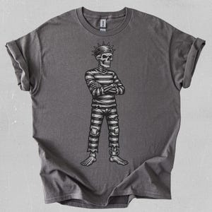 Puede incluir: Una camiseta gris con una ilustración en blanco y negro de un esqueleto con ropa de prisión. El esqueleto lleva una camisa y pantalones a rayas, con una cubierta para la cabeza con púas. El diseño está centrado en la camiseta.
