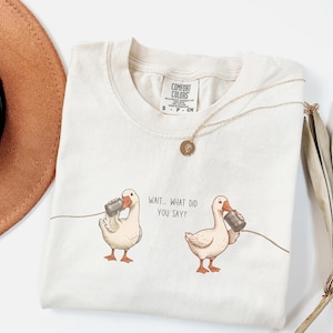 Puede incluir: Camiseta Comfort Colors color crema con una ilustración humorística de dos patos usando teléfonos de lata. El texto "Wait... What did you say?" está impreso en la camiseta. Un collar dorado con un colgante está sobre la camiseta.