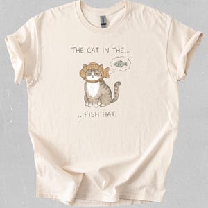 May include: Cream-colored t-shirt featuring a cartoon cat wearing a fish hat. The cat has a grumpy expression and a thought bubble with a fish. The text on the shirt reads "THE CAT IN THE... FISH HAT."
