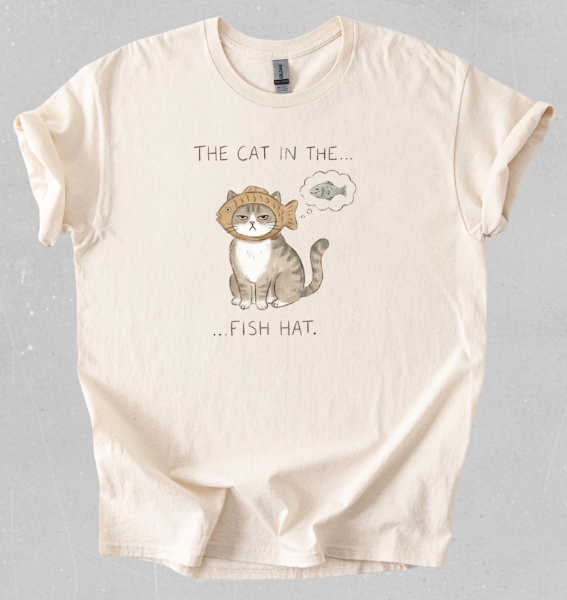 May include: Cream-colored t-shirt featuring a cartoon cat wearing a fish hat. The cat has a grumpy expression and a thought bubble with a fish. The text on the shirt reads "THE CAT IN THE... FISH HAT."