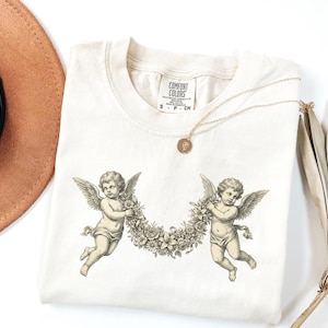 Puede incluir: Camiseta Comfort Colors color crema con una ilustración detallada de dos querubines sosteniendo una corona floral. Un collar de oro con un colgante descansa sobre la camisa. La camiseta tiene una etiqueta que dice "Comfort Colors".