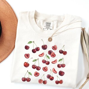 May include: Cream-colored t-shirt featuring a watercolor design of cherries. The design includes whole cherries, halved cherries, and cherry slices with green leaves. A gold necklace with a pendant is draped on the shirt.