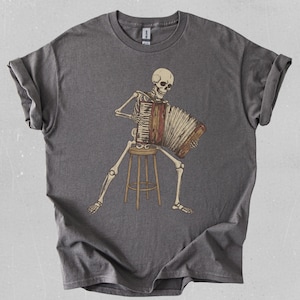 May include: A gray t-shirt featuring a skeleton playing an accordion while seated on a stool. The skeleton is depicted in a detailed, anatomical style, with the accordion in shades of brown and beige. The shirt is a comfortable, casual style.