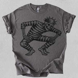 Puede incluir: Camiseta gris oscuro con una ilustración negra de un esqueleto con uniforme de prisionero. El esqueleto está agachado con las manos en las rodillas. El diseño presenta ropa a rayas y un tocado puntiagudo.