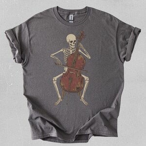 Puede incluir: Una camiseta gris con un esqueleto tocando el violonchelo. El esqueleto está sentado, sosteniendo el violonchelo con sus manos óseas. El violonchelo es de color marrón rojizo, y el diseño tiene un aspecto vintage y desgastado.