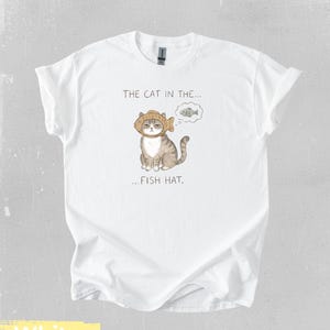 May include: White t-shirt featuring a whimsical illustration of a cat wearing a fish hat, with a thought bubble showing a fish. The text on the shirt reads "THE CAT IN THE... FISH HAT."