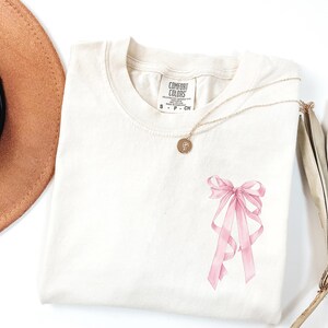 May include: Cream-colored t-shirt with a pink bow graphic. A gold necklace with a pendant rests on the shirt. The shirt has a tag that reads "Comfort Colors". A brown hat is visible in the upper left corner.