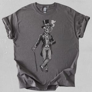 Puede incluir: Camiseta gris con una ilustración en blanco y negro de un esqueleto con traje y sombrero de copa, fumando un cigarrillo. El esqueleto sostiene un bastón. El diseño es detallado y formal.