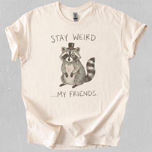 Puede incluir: Camiseta color crema con una ilustración caprichosa de un mapache con sombrero de copa y monóculo. El texto "STAY WEIRD...MY FRIENDS" está impreso encima y debajo del mapache. La camiseta tiene cuello redondo y mangas cortas.