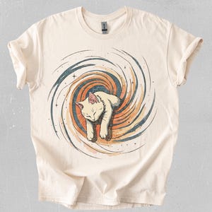 Puede incluir: Camiseta de color crema con una ilustración de un gato blanco dentro de un diseño de galaxia en espiral. El gato está representado durmiendo, con el diseño de la galaxia en tonos naranja, azul y marrón. La camiseta tiene mangas cortas y cuello redondo.