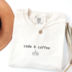 Puede incluir: Camiseta color crema con las palabras "code & coffee" en negro y un icono de taza de café. Un collar dorado y un sombrero marrón son parcialmente visibles en la imagen.
