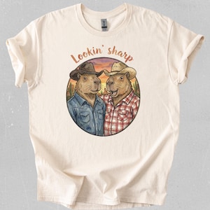 Puede incluir: Camiseta color crema con un gráfico circular de dos carpinchos con sombreros y camisas de vaquero, sobre un fondo de puesta de sol desértica. El texto "Lookin' sharp" está arqueado sobre el gráfico en marrón.