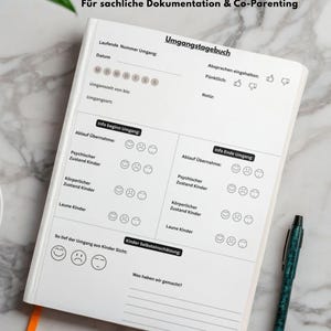 Peut inclure: Un journal blanc ouvert, intitulé "Umgangstagebuch" et sous-titré "Für sachliche Dokumentation & Co-Parenting", est posé sur une surface en marbre. Le journal contient des sections pour la documentation et un stylo est à droite.