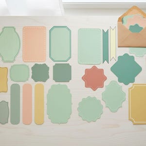 May include: A collection of pastel-colored paper ephemera, including labels, tags, and decorative shapes. The assortment features various shapes, including rectangles, banners, and scalloped edges. A kraft paper envelope and a pair of scissors are also present.