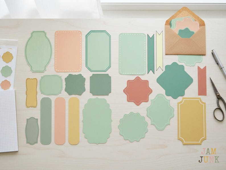 May include: A collection of pastel-colored paper ephemera, including labels, tags, and decorative shapes. The assortment features various shapes, including rectangles, banners, and scalloped edges. A kraft paper envelope and a pair of scissors are also present.