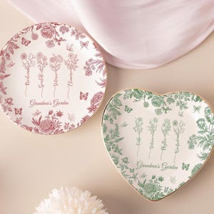 Puede incluir: Dos platos decorativos con diseños florales. Uno es redondo con un patrón floral rojo y blanco, el otro tiene forma de corazón con un patrón floral verde y blanco. Ambos platos tienen bordes dorados y el texto "Grandma's Garden".