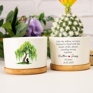 Może przedstawiać: Dwie białe ceramiczne donice z bambusowymi spodkami. Jedna przedstawia ilustrację płaczącej wierzby, druga ma tekst: „Jak wierzba... razem. Kelton & Jessy 10 lat 10.22.2025.” W jednej znajduje się kaktus z pomarańczowym kwiatem.