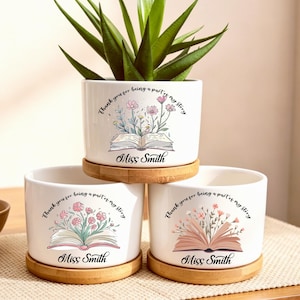 Può includere: Tre vasi cilindrici in ceramica bianca con sottovasi in bambù. Ogni vaso presenta un disegno di un libro aperto con fiori che emergono dalle pagine e il testo "Thank you for being a part of my story" e "Miss Smith" in caratteri corsivi.