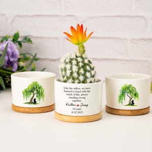 May include: Three white ceramic plant pots with bamboo bases. One holds a cactus with an orange and yellow flower. The other two feature a willow tree design and text that reads "Like the willow... Kelton & Jessy 10 years 12.22.2025."