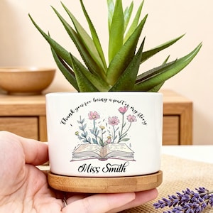May include: A white ceramic planter with a bamboo base containing a green succulent. The planter features a graphic of an open book with flowers growing from the pages and the text "Thank you for being a part of my story" and "Miss Smith".