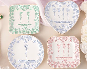 Puede incluir: Cuatro platos decorativos de cerámica en formas cuadradas, de corazón y redondas. Cada plato presenta diseños florales y texto en varios colores, incluyendo verde, azul y rosa. Los platos tienen bordes dorados.