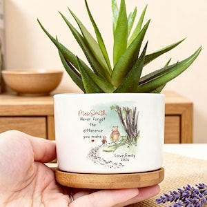 May include: A white ceramic cylindrical planter with a bamboo saucer containing a green succulent. The planter features a printed illustration of Winnie the Pooh and Piglet walking on a path with the text: Mrs Smith, Never forget the difference you make. Love, Emily 2026.