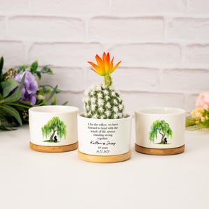 May include: Three white ceramic planters with bamboo bases. One planter holds a cactus with an orange and yellow flower. The other two planters feature a watercolor willow tree design. Text on one planter reads: "Like the willow...Kelton & Jessy 10 years 1.22.2025."