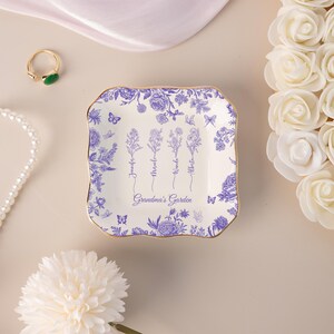 Pu&ograve; includere: Un vassoio quadrato in ceramica bianca con bordo dorato e design floreale blu. Il piatto &egrave; decorato con fiori, farfalle e le parole "Grandma's Garden" e nomi. Un anello e una collana di perle sono sullo sfondo.