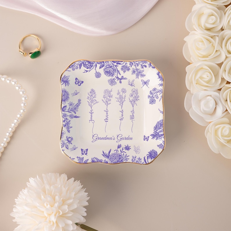 Pu&ograve; includere: Un vassoio quadrato in ceramica bianca con bordo dorato e design floreale blu. Il piatto &egrave; decorato con fiori, farfalle e le parole "Grandma's Garden" e nomi. Un anello e una collana di perle sono sullo sfondo.