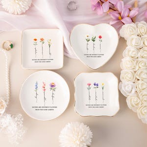 Puede incluir: Cuatro platos de cerámica blanca para baratijas en formas cuadrada, de corazón, redonda y festoneada. Cada plato presenta un diseño floral, borde dorado y el texto "SISTERS ARE DIFFERENT FLOWERS FROM THE SAME GARDEN".