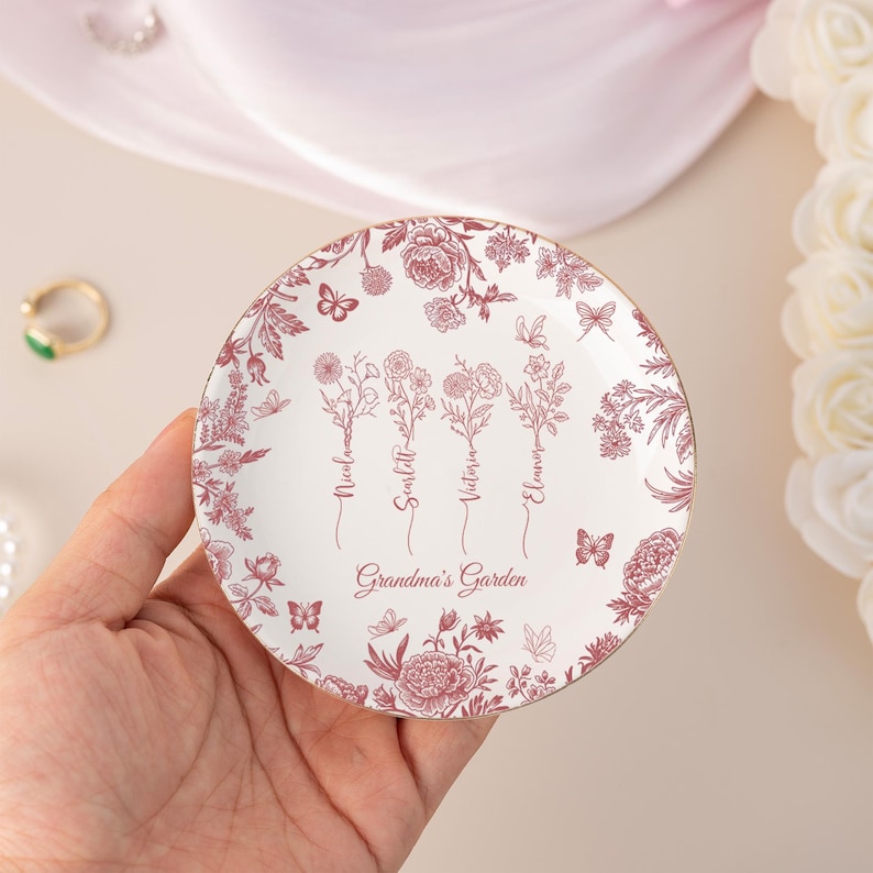Pu&ograve; includere: Un piatto portagioie in ceramica bianca con un motivo floreale bordeaux. Il piatto presenta le parole "Grandma's Garden" e nomi. Il bordo &egrave; dorato. Farfalle e fiori fanno parte del design.