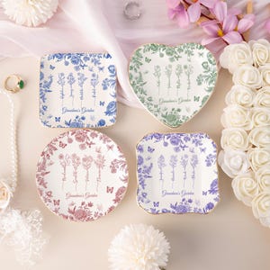 Pu&ograve; includere: Quattro piatti decorativi in ceramica di varie forme: quadrata, a cuore e rotonda. Ogni piatto presenta disegni floreali blu, verdi e rossi, con la scritta "Grandma's Garden". Farfalle e bordi dorati.