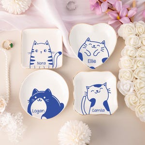 以下が含まれることがあります： 金色の縁取りが施された4つの白いセラミック製の小物入れ。それぞれに青い猫のデザインと名前（Nora、Ellie、Logan、Camila）が描かれています。正方形、ハート型、丸型、スカラップ型です。